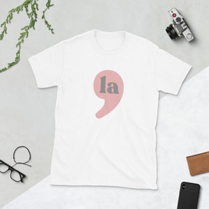 Puede incluir: Camiseta blanca con una coma rosa y la palabra "la" en gris.