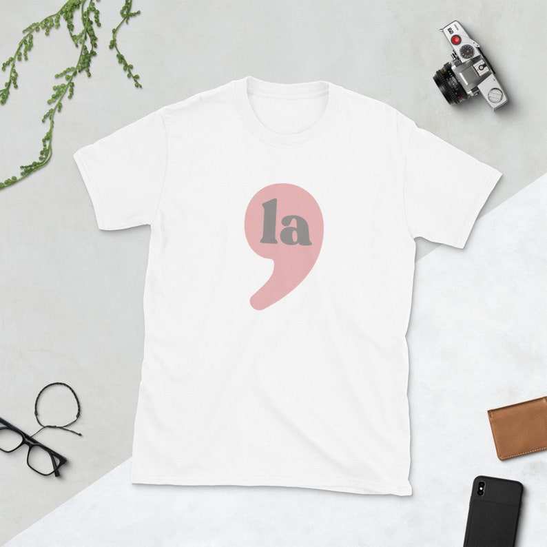 Puede incluir: Camiseta blanca con una coma rosa y la palabra "la" en gris.