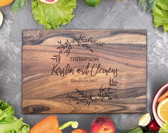 May include: A rectangular wooden cutting board with a dark brown wood grain pattern. The center features an engraved laurel wreath design surrounding the text "THOMPSON", "Kerstin and Clemens", and the date "February 6, 2017" in a mix of fonts.
