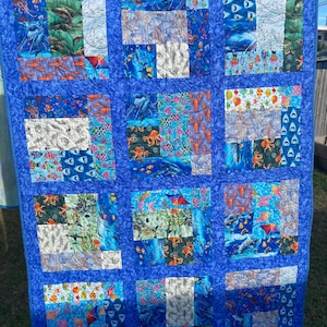 May include: A blue and white patchwork quilt with a nautical theme. The quilt features various sea creatures, including fish, sharks, and an octopus. The quilt is made of different fabrics with various patterns, including stripes, polka dots, and floral prints.