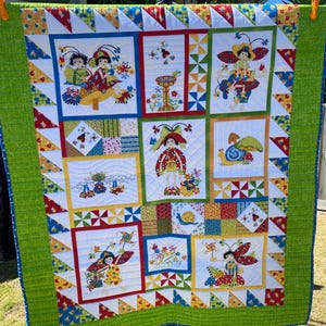 Könnte beinhalten: Eine farbenfrohe Quiltdecke mit grünem Rand und weißem Hintergrund. Die Decke zeigt verschiedene fantasievolle Motive, darunter Schmetterlinge, Marienkäfer und Blumen. Die Decke besteht aus Quadraten und Dreiecken, und die Motive sind im Patchwork-Stil zusammengenäht.