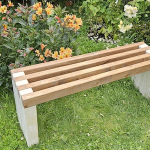 May include: A wooden garden bench with a slatted seat and light-coloured legs. The bench is made of natural wood and is set in a garden with green grass and colourful flowers. The bench is approximately 122 cm long.