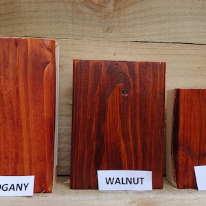 May include: Three wooden blocks stained in different shades of brown. The blocks are labeled "Mahogany", "Walnut", and "Teak" on white labels. The wood grain is visible, and the blocks are arranged against a wooden background.