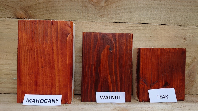 May include: Three wooden blocks stained in different shades of brown. The blocks are labeled "Mahogany", "Walnut", and "Teak" on white labels. The wood grain is visible, and the blocks are arranged against a wooden background.