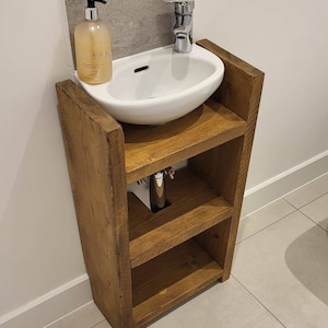 May include: A rustic, wooden vanity with a white, oval sink and chrome faucet. The vanity has two shelves and is mounted against a gray tiled wall. A bottle of liquid soap sits on the counter. The vanity is made of reclaimed wood.