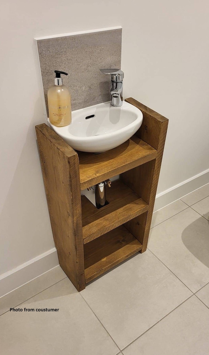 May include: A rustic, wooden vanity with a white, oval sink and chrome faucet. The vanity has two shelves and is mounted against a gray tiled wall. A bottle of liquid soap sits on the counter. The vanity is made of reclaimed wood.