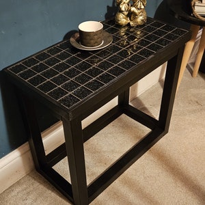 May include: Black side table with a black and gold mosaic tile top. The table has a single shelf and a square base. Two gold elephant figurines are on the table.