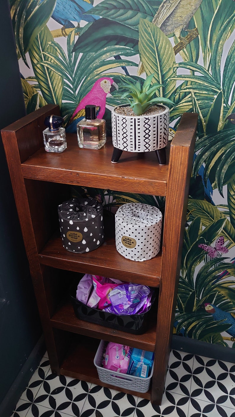 May include: A dark brown wooden shelf unit with various items. Includes a small potted plant, perfume bottles, and rolls of toilet paper. The toilet paper rolls are wrapped in patterned paper. The bottom shelves hold baskets of feminine hygiene products.