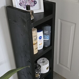 May include: A black wooden shelf unit with four shelves. The top shelf holds a framed artwork with a button design, and a bottle of perfume. The shelves below hold toiletries and storage containers. A small white basket is on the bottom shelf.