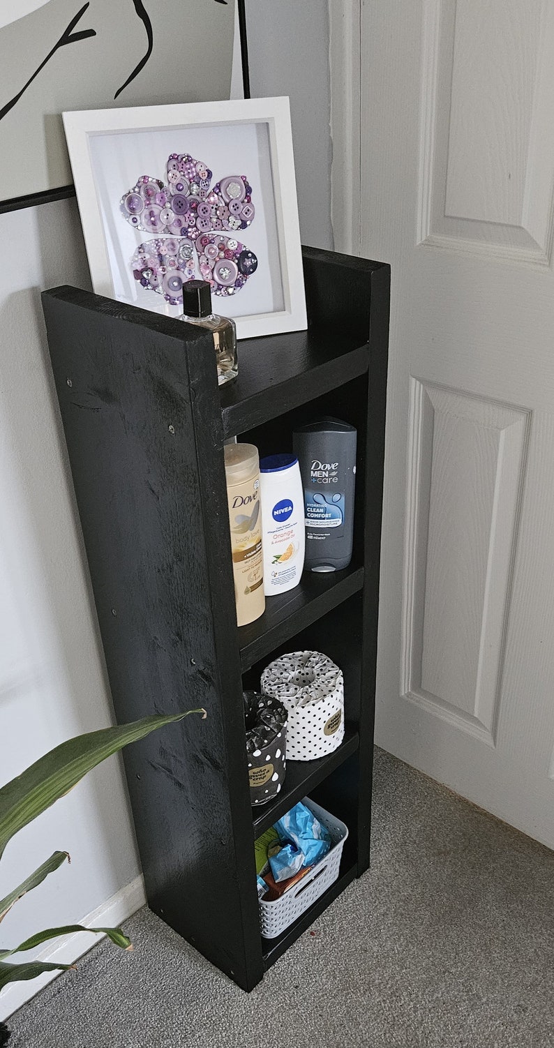 May include: A black wooden shelf unit with four shelves. The top shelf holds a framed artwork with a button design, and a bottle of perfume. The shelves below hold toiletries and storage containers. A small white basket is on the bottom shelf.