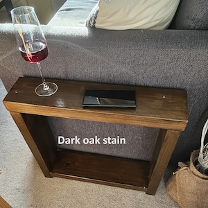 May include: A dark oak stained wooden side table with a shelf. The table has a rectangular top and is sitting next to a grey sofa. A mobile phone is on the top of the table. "Dark oak stain" is written on the table.