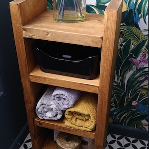 May include: Wooden bathroom shelf unit with three shelves. The top shelf holds a glass diffuser with reeds. The middle shelf has a black plastic container. Rolled gray and yellow towels are on the lower shelves. The unit is against a tropical wallpaper.