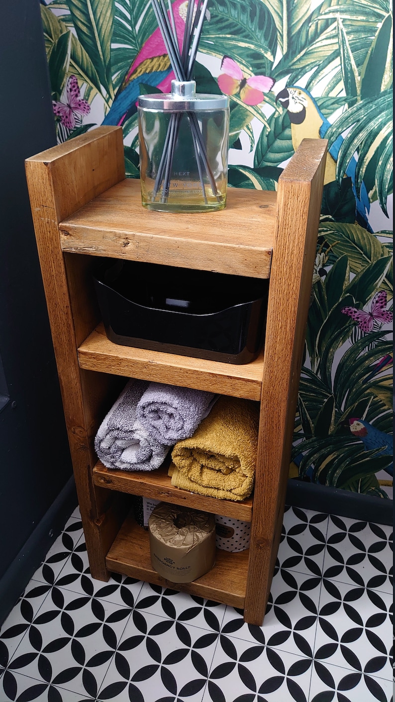 May include: Wooden bathroom shelf unit with three shelves. The top shelf holds a glass diffuser with reeds. The middle shelf has a black plastic container. Rolled gray and yellow towels are on the lower shelves. The unit is against a tropical wallpaper.