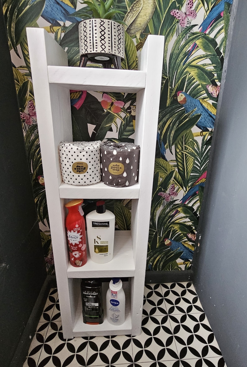 May include: A white wooden shelf unit with four shelves, holding various bathroom products. The top shelf has a small potted plant. The second shelf has two rolls of toilet paper. The third shelf has a red spray bottle and a white shampoo bottle. The bottom shelf has two bottles.