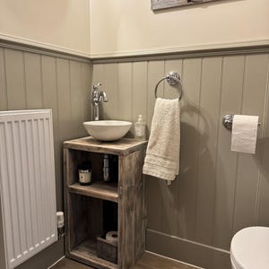 May include: A rustic wooden vanity with a white ceramic sink and chrome faucet. The vanity has open shelving and a woven basket. A white towel hangs on a chrome ring, and a roll of toilet paper is mounted on the wall. The bathroom has a neutral color scheme.