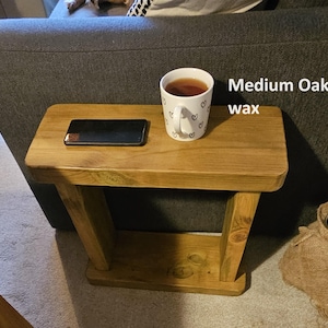 May include: A small wooden side table with a cup of tea and a mobile phone on top. The table is made of medium oak wood and has a natural finish. The text "Medium Oak wax" is visible in the image.