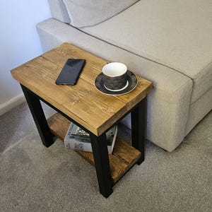 May include: A small, rustic side table with a wooden top and a lower shelf. The table has black metal legs and a natural wood finish. A smartphone and a teacup and saucer are on the table. Books are on the lower shelf.