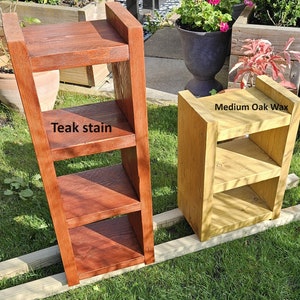 May include: Two wooden shelves, one stained with teak and the other with medium oak wax. The teak shelf is a leaning ladder design with three shelves. The oak shelf is a small, boxy design with three shelves. Both are outdoors.