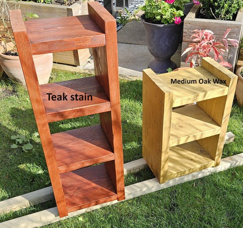 May include: Two wooden shelves, one stained with teak and the other with medium oak wax. The teak shelf is a leaning ladder design with three shelves. The oak shelf is a small, boxy design with three shelves. Both are outdoors.