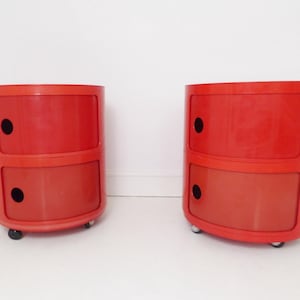 Op de afbeelding: Twee rode, cilindrische nachtkastjes met elk twee gestapelde compartimenten. De kasten hebben zwarte ronde handgrepen en kleine wieltjes. Het afgeronde ontwerp en de heldere kleur geven ze een retro-esthetiek.