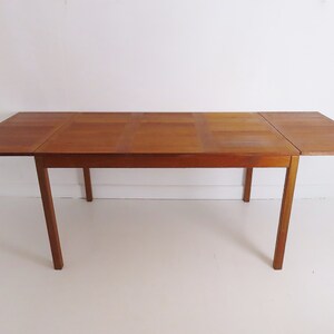 May include: A rectangular wooden dining table with a warm brown finish. The table has a central section and two drop-leaf extensions. The legs are straight and angled slightly outward. The table is approximately 152 cm long when fully extended.