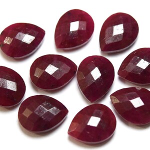 May include: A collection of faceted, teardrop-shaped gemstones in a deep red hue. The gems are arranged on a white surface, showcasing their polished surfaces and internal reflections. The gemstones are likely rubies.