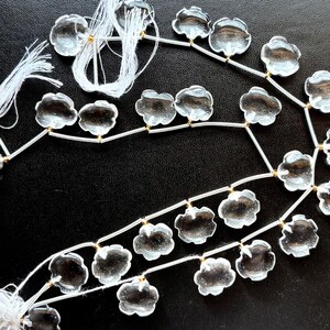 Puede incluir: Una hebra de cuentas de cristal transparente en forma de flor con alambre plateado y detalles dorados. Las cuentas están enhebradas con hilo blanco.