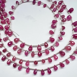 May include: Strands of faceted, teardrop-shaped ruby gemstones in varying shades of pink and red. The gemstones are strung on thin, silver-colored wire, creating a layered effect. The beads are arranged on a white background.