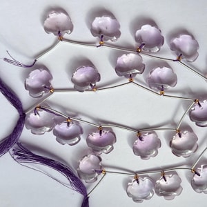 Puede incluir: Una hebra de cuentas de piedras preciosas de color púrpura claro en forma de flor con acentos plateados y dorados. Las cuentas están enhebradas en un alambre plateado con borlas moradas en cada extremo.