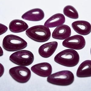 May include: A collection of 18 teardrop-shaped, faceted, purple gemstones. The gemstones are arranged on a white surface.