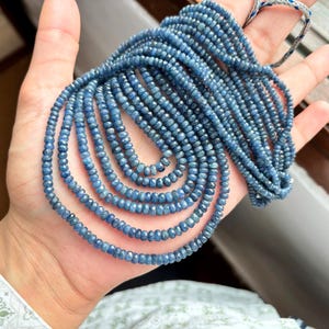 Può includere: Un filo di perle di zaffiro blu, sfaccettate e rotonde, infilate su un cordoncino. Le perle hanno un diametro di circa 3 mm e sono disposte in un motivo sciolto e fluido.