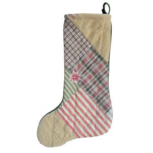 May include: A quilted Christmas stocking with a patchwork design featuring plaid, striped, and solid fabric in shades of green, red, and beige. The stocking has a green felt cuff and a small embroidered flower.