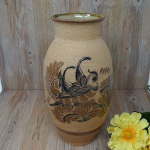 May include: A beige ceramic vase with a bird and floral design. The vase has a wide body and a narrow neck. The bird is black and brown, and the flowers are brown and green. The vase is approximately 12 inches tall.