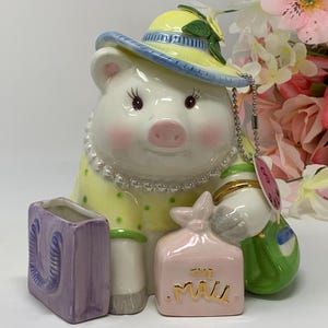 May include: A ceramic pig figurine wearing a yellow hat and dress with green polka dots, a pearl necklace, and holding a green purse. It has a pink shopping bag that says "THE MALL" and a purple shopping bag.