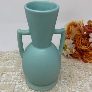Puede incluir: Un jarrón de cerámica azul claro con acabado mate y sutiles motas oscuras. El jarrón presenta un cuerpo redondeado, un cuello acampanado y dos asas simétricas a los lados. La superficie tiene una textura suave y un color uniforme.