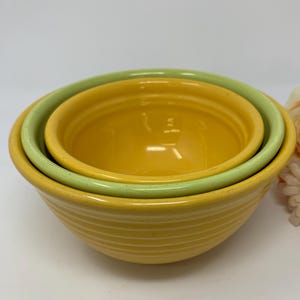 May include: A trio of ceramic mixing bowls in yellow and green hues. The largest bowl is a vibrant yellow with a ribbed design, with two smaller bowls nested inside. A decorative flower is in the background.