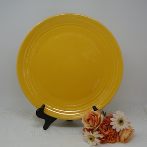 Könnte beinhalten: Ein hellgelber Teller aus Keramik mit leicht erhöhtem Rand. Der Teller steht auf einem weißen Ständer.