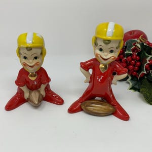 Könnte beinhalten: Zwei Keramikfiguren von Footballspielern. Einer kniet und hält einen Football, der andere steht mit den Händen in den Hüften. Beide tragen rote Uniformen und gelbe Helme mit weißen Streifen. Dekorative rote Beeren und Äpfel sind im Hintergrund.
