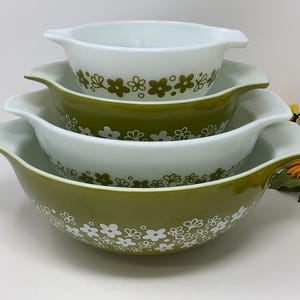 Può includere: Un set di tre ciotole impilabili in vetro opalino con manici. Le ciotole presentano un motivo floreale ripetuto in bianco e verde oliva. La ciotola più piccola è bianca con fiori verdi, mentre le due più grandi sono verde oliva con fiori bianchi.