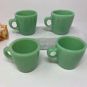 May include: A set of four matching light green glass mugs with a smooth, opaque finish and a rounded handle. Each mug features a wide, open top that tapers down toward a smaller base, creating a simple, functional shape for holding beverages.