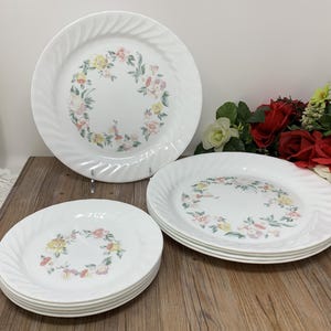 Pode incluir: Pratos de cerâmica brancos com um desenho de coroa floral em rosa, amarelo e verde. Os pratos têm uma borda em espiral. Uma pilha de pratos e pratos individuais são exibidos em uma superfície de madeira.