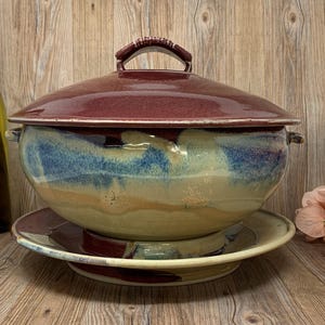 Puede incluir: Una sopera de cerámica con tapa, apoyada sobre un plato a juego. La sopera y la tapa son de color burdeos oscuro, mientras que el cuenco tiene tonos azules, beiges y blancos. El asa de la tapa es de color burdeos.