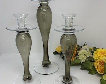 Peut inclure: Un ensemble de trois bougeoirs en verre avec des tiges en forme de goutte d'eau de couleur fumée et des bases circulaires transparentes. Chaque bougeoir possède une coupelle transparente large et plate pour la cire.