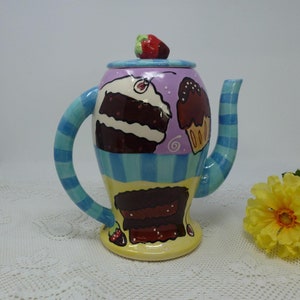 May include: A ceramic teapot with a blue and white striped handle and a colorful design of cakes and cupcakes. The teapot has a purple lid with a small red strawberry on top.