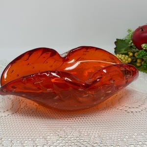 Puede incluir: Un cenicero de vidrio naranja brillante con una superficie texturizada y con burbujas. El cenicero tiene la forma de una boca grande abierta.