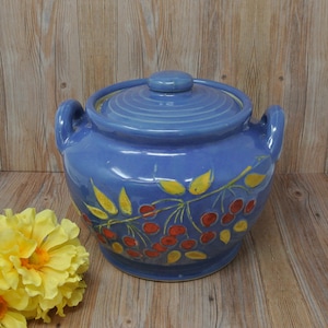 May include: A blue ceramic crock with a lid, decorated with a yellow and red floral design. The crock has two handles on the sides.