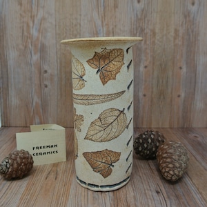 May include: A white ceramic vase with a brown leaf design. The vase is tall and cylindrical with a rounded top. The leaves are arranged in a vertical pattern around the vase.