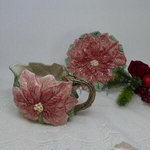 Puede incluir: Un juego de jarra para crema y plato a juego de cerámica con motivos de flor de Pascua. La jarra tiene un asa de rama marrón y está adornada con una gran flor de Pascua rosa y hojas verdes. El plato tiene forma de flor de Pascua.