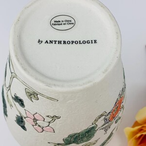 May include: Off-white ceramic vase with floral design in pink, orange, and green. The vase has a textured surface and the words "by ANTHROPOLOGIE" are printed on the bottom. The vase is marked "Made in China".