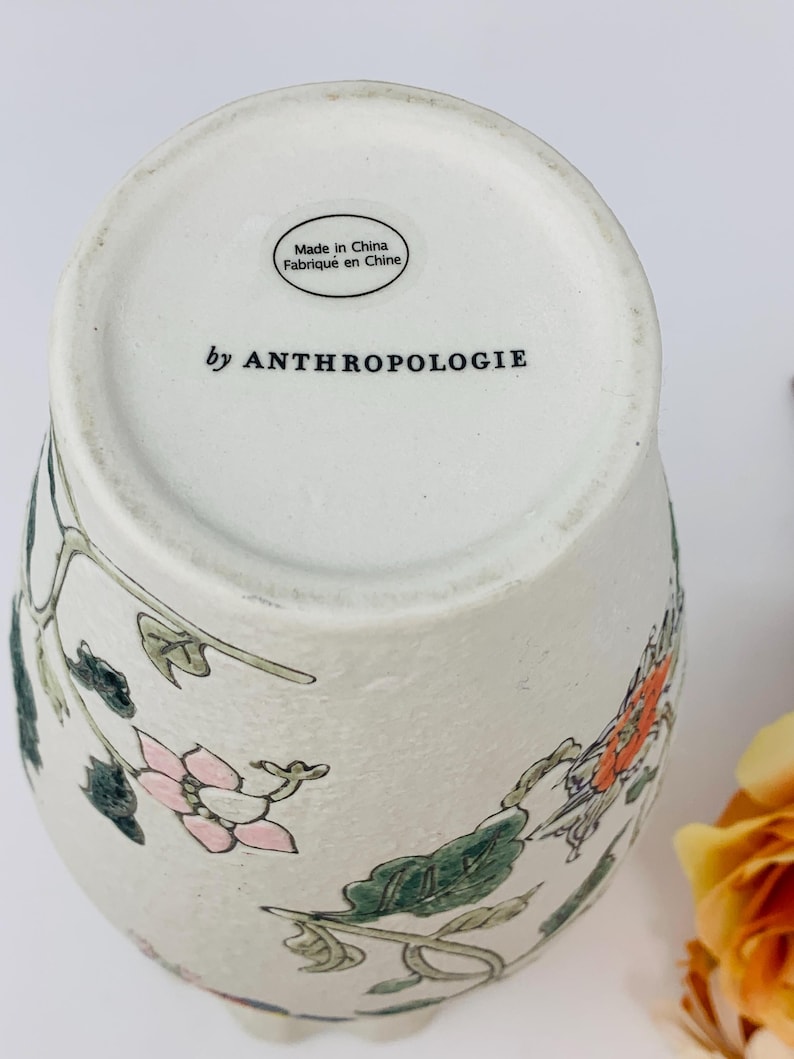 May include: Off-white ceramic vase with floral design in pink, orange, and green. The vase has a textured surface and the words "by ANTHROPOLOGIE" are printed on the bottom. The vase is marked "Made in China".